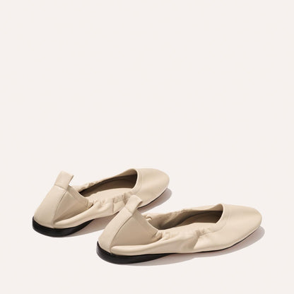 The Pascale - Bone Nappa ballet flats by Margaux, featuring beige nappa leather, elasticized edges, and black soles, are displayed on a light background—one shoe facing forward, the other slightly behind and to the side.