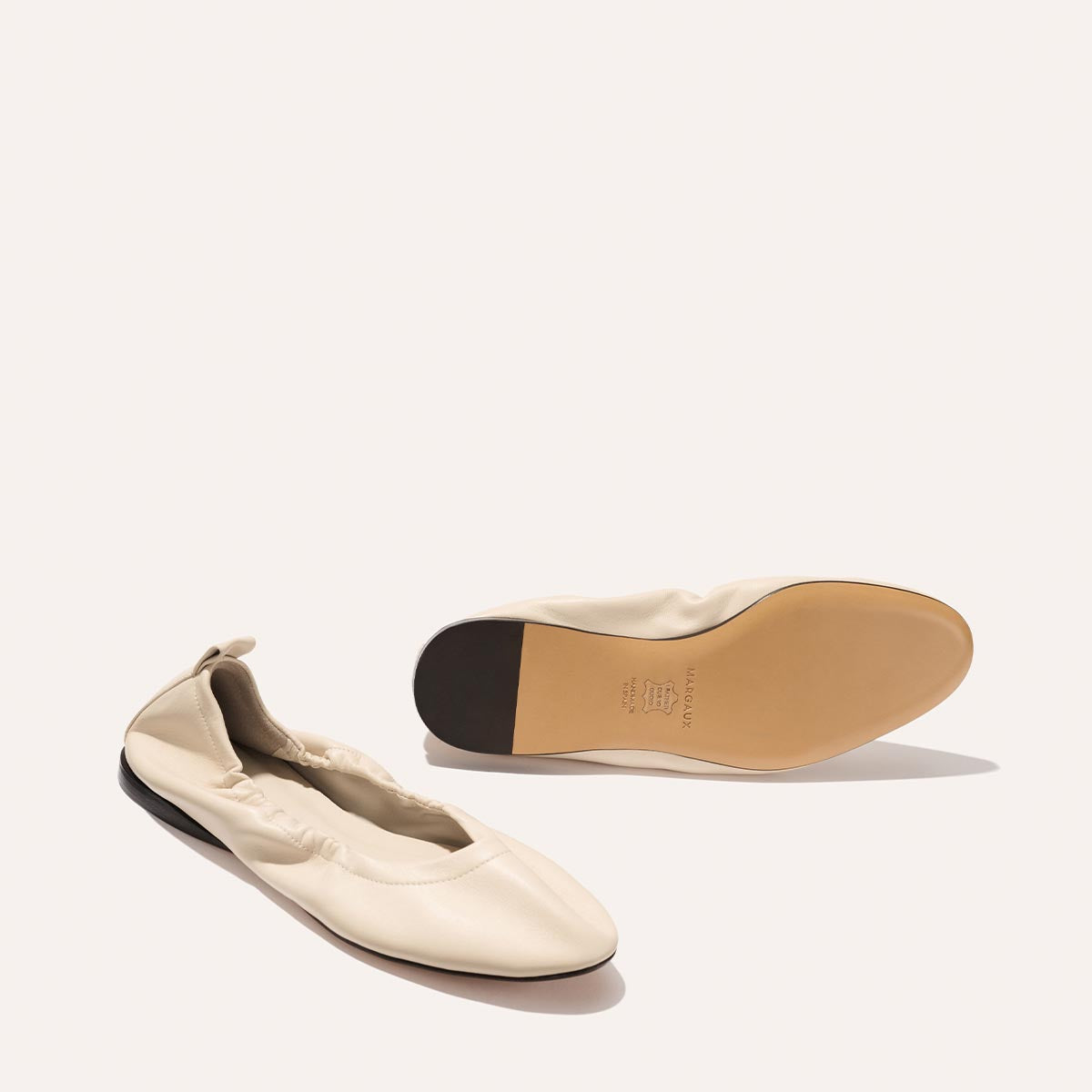 The Pascale - Bone Nappa by Margaux: cream-colored ballet flats with elastic sides and tan soles. Displayed on a light background, one shoe is upright while the other reveals its tan sole.