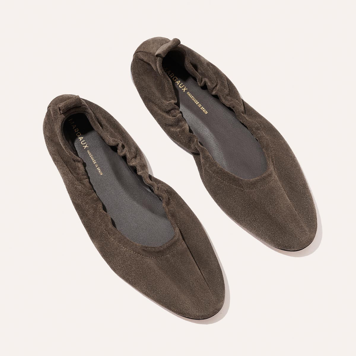 A pair of Margaux's The Pascale ballet flats in English Brown Suede, featuring ruched edges, an elasticated back, pointed almond-shaped toes, and dark inner soles, are placed side by side on a light background.