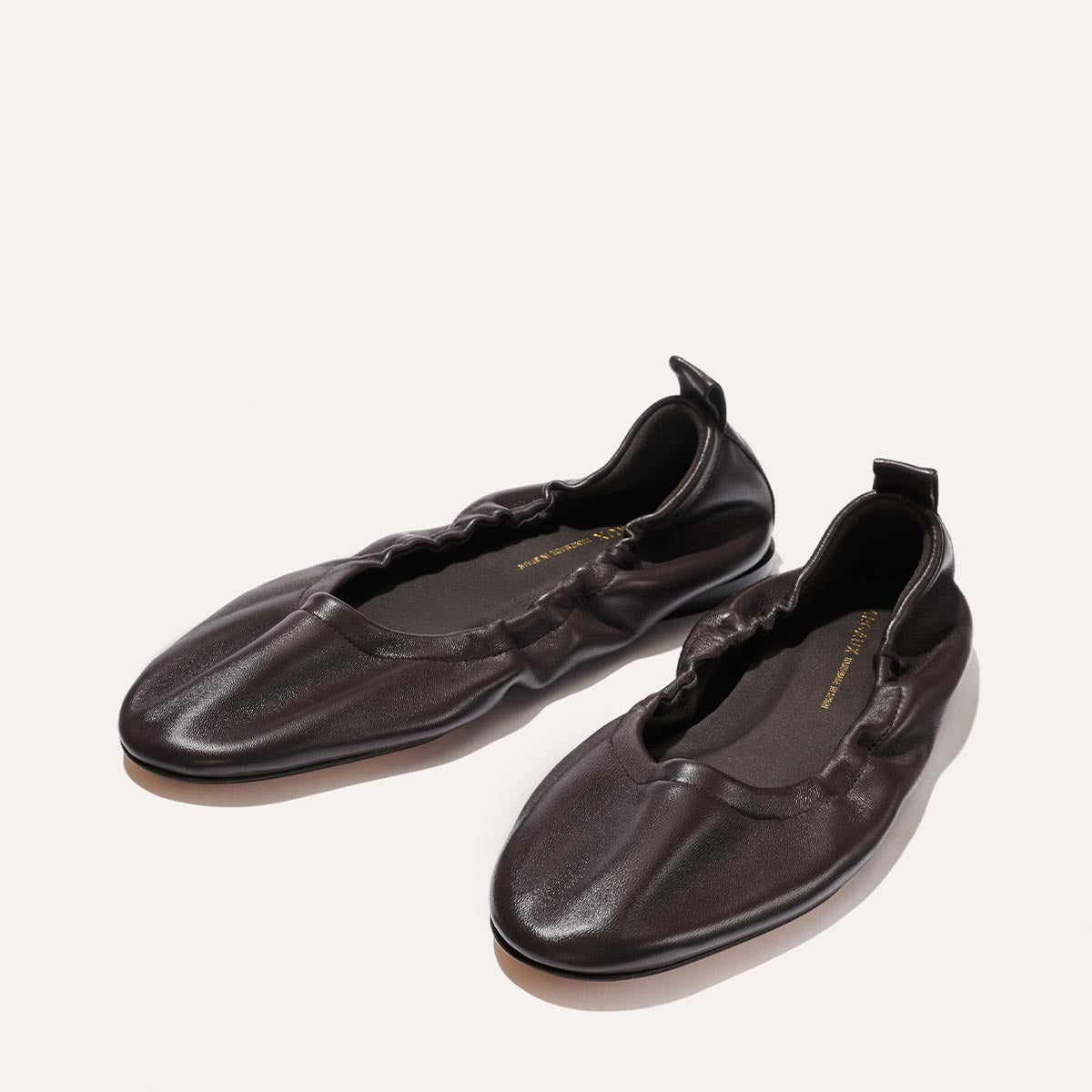 A pair of The Pascale - Espresso Nappa slip-on ballet flats by Margaux, crafted from supple dark brown leather with ruched detailing and an elasticated back pull tab, displayed on a light background.