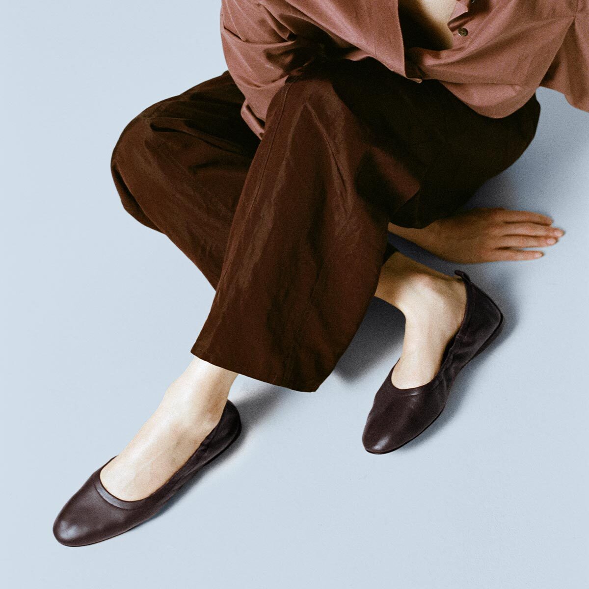 A person sits cross-legged on the floor against a light background, head out of frame, wearing dark loose pants and a mauve shirt. They are showcasing Margaux’s The Pascale - Espresso Nappa: supple leather deconstructed ballet flats with an elasticated back.