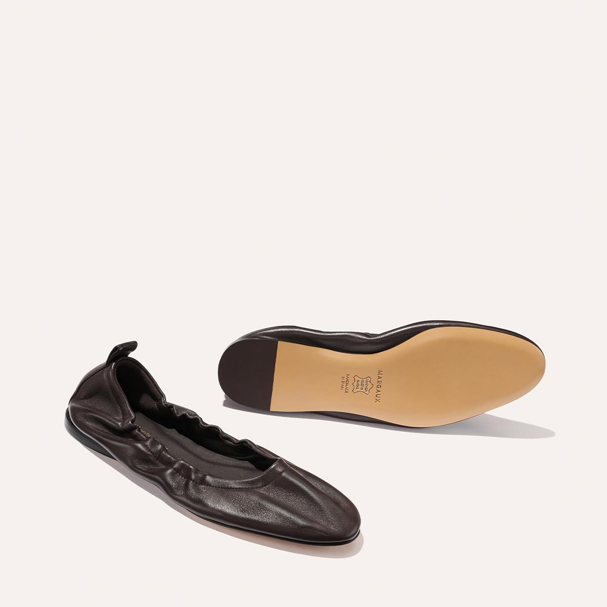 A pair of Margaux's "The Pascale - Espresso Nappa" ballet flats in dark brown supple leather, featuring an elasticated back for a comfortable fit—one shoe upright and the other showing the tan branded sole, displayed on a light background.