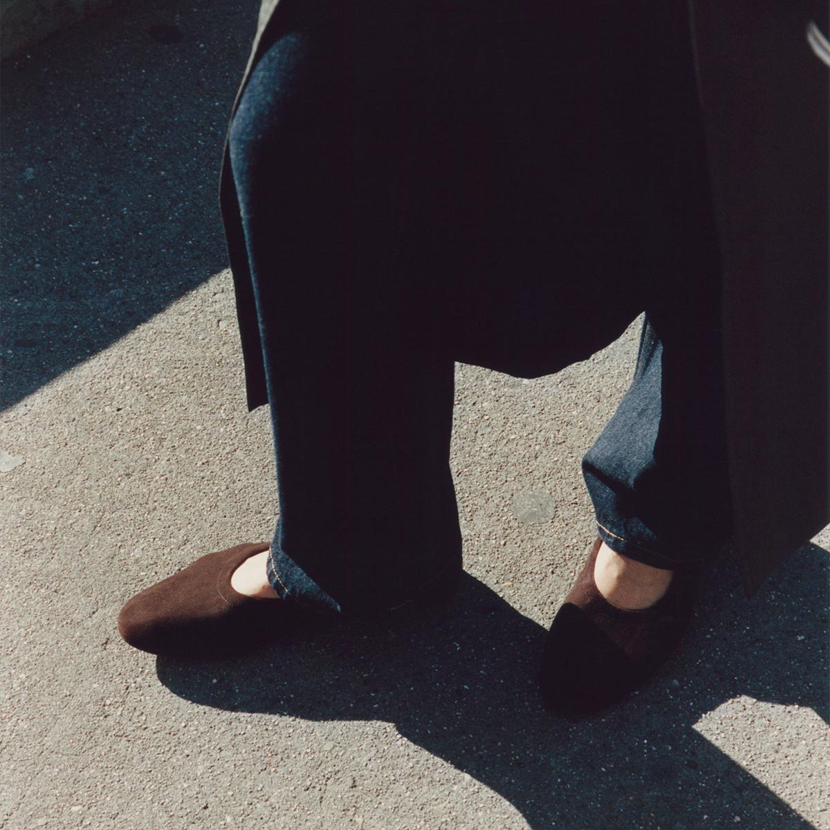 On a sunlit sidewalk, someone in dark pants wears Margaux’s The Pascale – Espresso Suede ballet flats, mixing brown and black for a playful contrast. Soft suede shoes cast shadows on the concrete; only the lower half of the outfit is shown.