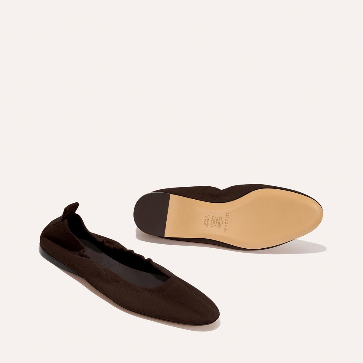 The Pascale - Espresso Suede by Margaux: A pair of dark brown ballet flats, one resting flat and the other on its side to reveal the light tan sole and small heel pull tab. These shoes feature a simple, elegant design.