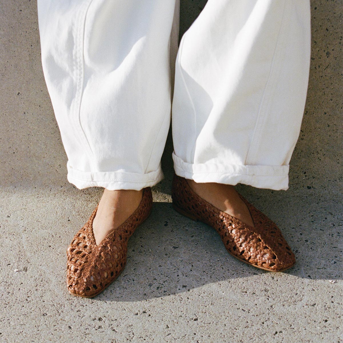 A person wearing high-rise loose white pants and Margaux’s The Woven Paz - Saddle Leather flats stands on textured concrete, with sunlight casting soft shadows.