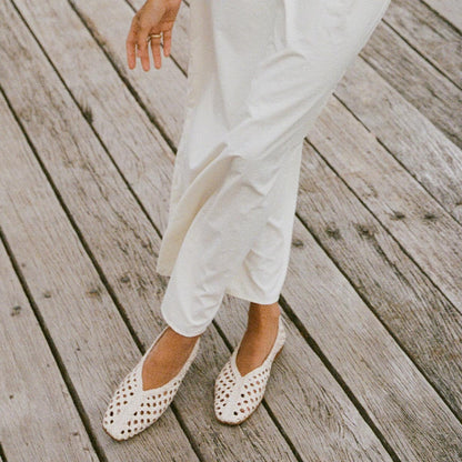 A person wearing loose white pants and Margauxs The Woven Paz - Ecru Leather slip-on shoes stands on a wooden deck, with only the lower half of their body and a hand visible.