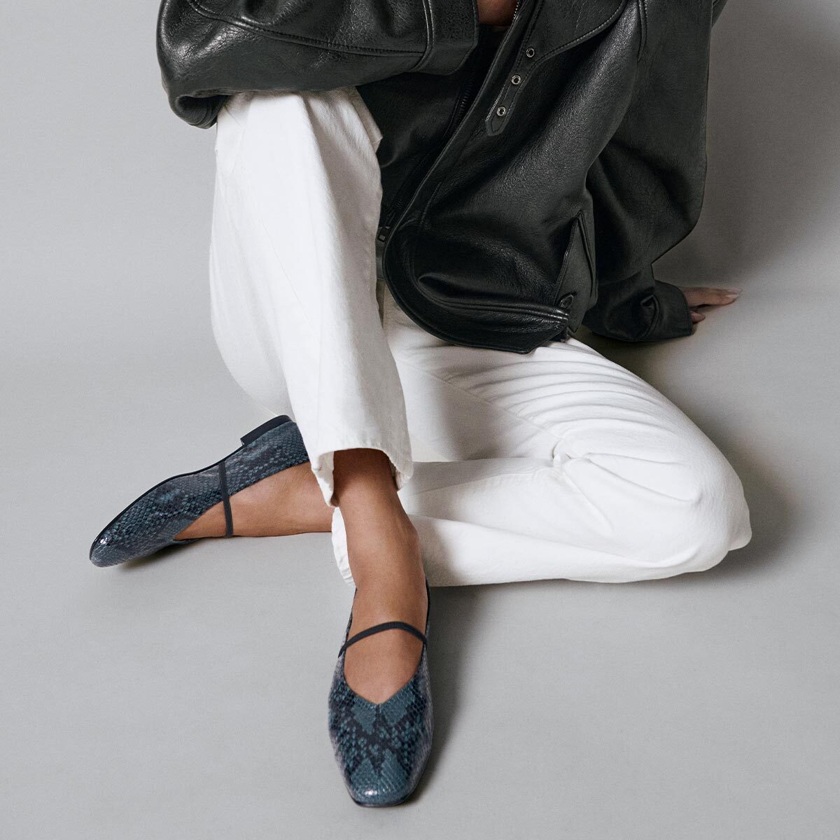 A person sits on the floor wearing a black leather jacket, white pants, and Margaux’s The Phoebe flats in Aqua Python Embossed. These blue, snakeskin-patterned Mary Jane shoes are handmade using sacchetto construction for an exceptionally flexible fit and are crafted to be the softest flats you’ll ever wear. The person’s face is not visible.