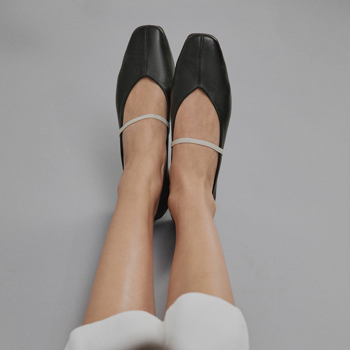 Wearing The Phoebe - Black and Ivory Nappa flats by margaux-development, a person with legs extended sits on a light gray surface. The shoes are soft, black, square-toed with thin, ivory-colored straps.