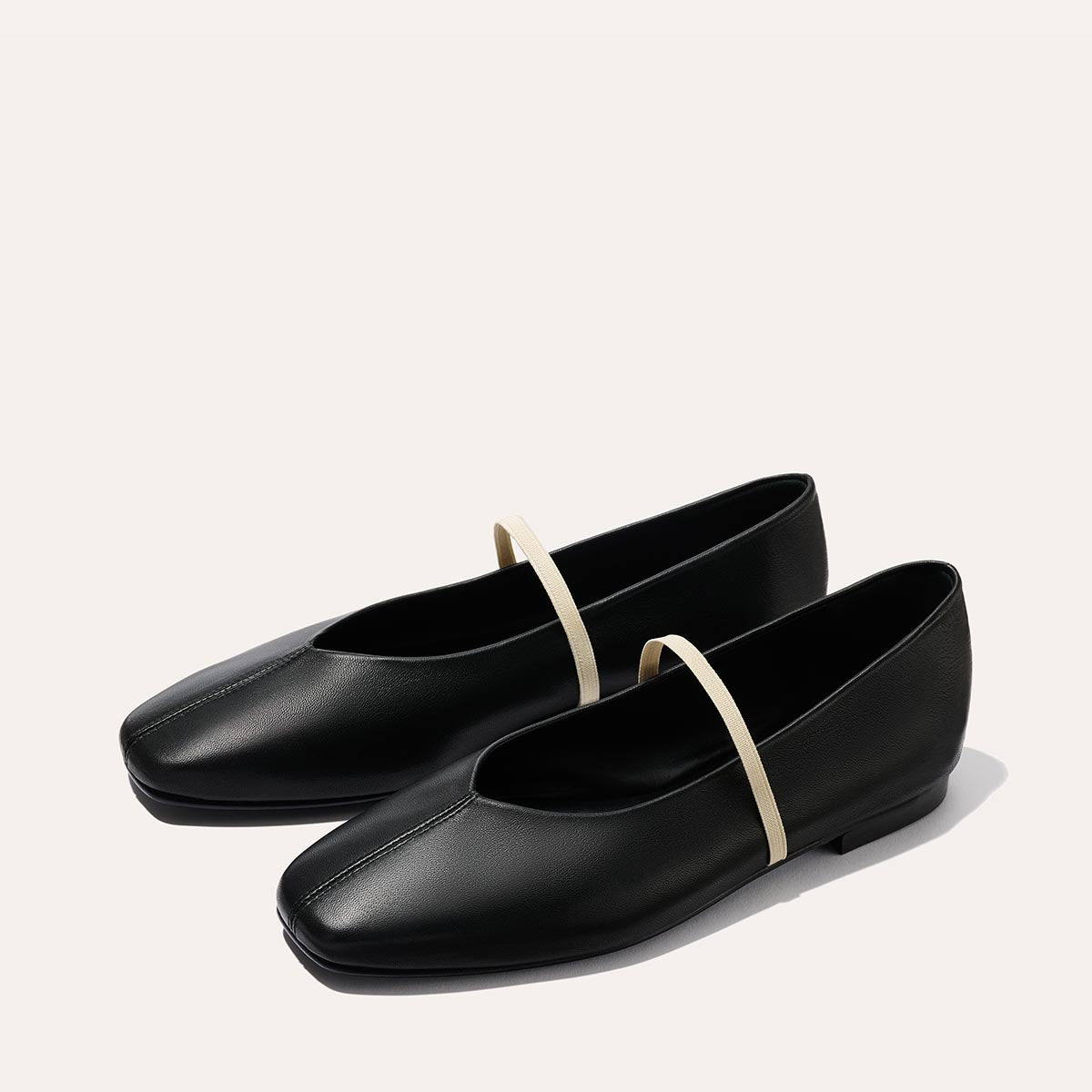 The Phoebe - Black and Ivory Nappa by margaux-development: handmade black ballet shoes with beige elastic straps, minimalist design, and sacchetto construction, shown on a plain light background.