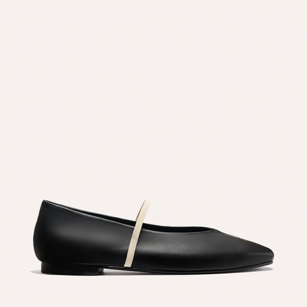 The Phoebe - Black and Ivory Nappa by margaux-development is a black ballet flat with a rounded toe and thin beige strap across the top, shown in a side view against a plain light background. Crafted using sacchetto construction.