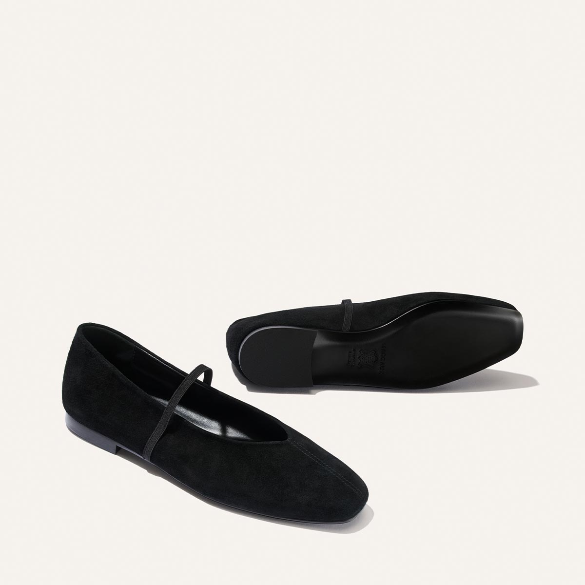 Two “The Phoebe - Black Suede” ballet flats by Margaux, one standing upright and the other positioned on its side to reveal the sole. These minimalist black suede shoes feature a thin strap across the top and are displayed against a plain, light background.