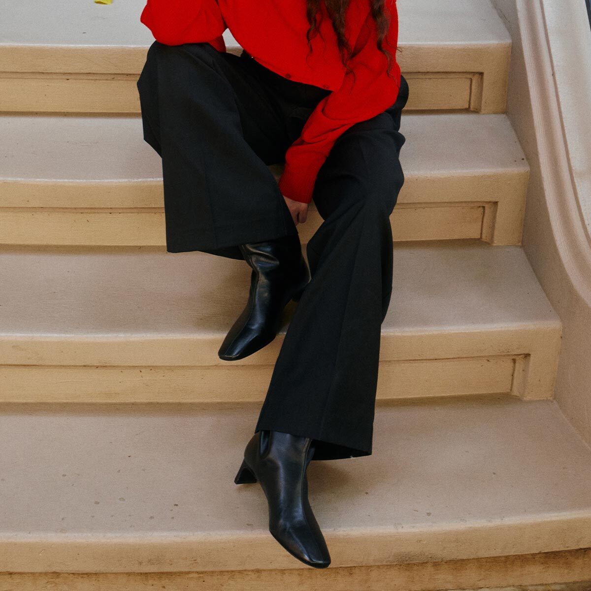 A person sits on stairs wearing a red long-sleeve top, black wide-leg pants, and Margaux’s The Brera Boot in Black Nappa. Their face is not visible in the image.