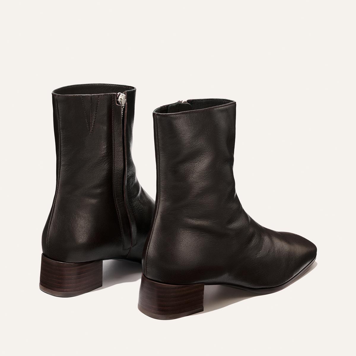 A pair of Margaux’s The Phoebe Boot in Dark Chocolate Calf leather, featuring side zippers, a low stacked leather heel, and a modern square toe, shown from the back and side against a plain light background.
