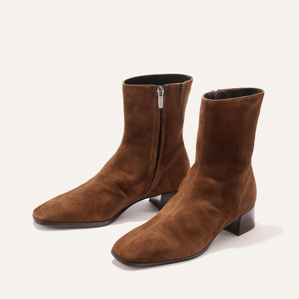 Introducing The Phoebe Boot - Ganache Suede by Margaux: a pair of brown suede ankle boots with side zippers and low block heels, expertly crafted using the artisanal sacchetto technique for outstanding flexibility and comfort. Featuring a padded insole, these boots are showcased against a light background.