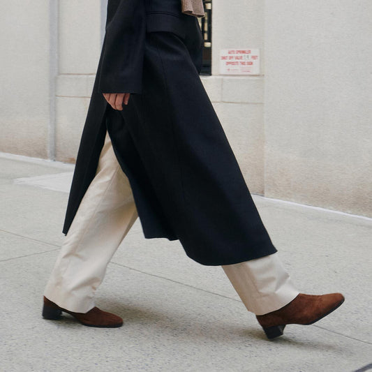 A person walking on a sidewalk, wearing cream-colored pants, Margaux’s The Phoebe Boot in Ganache Suede, and a long dark coat. Only the lower half of their body is visible. A white wall and a small sign are in the background.