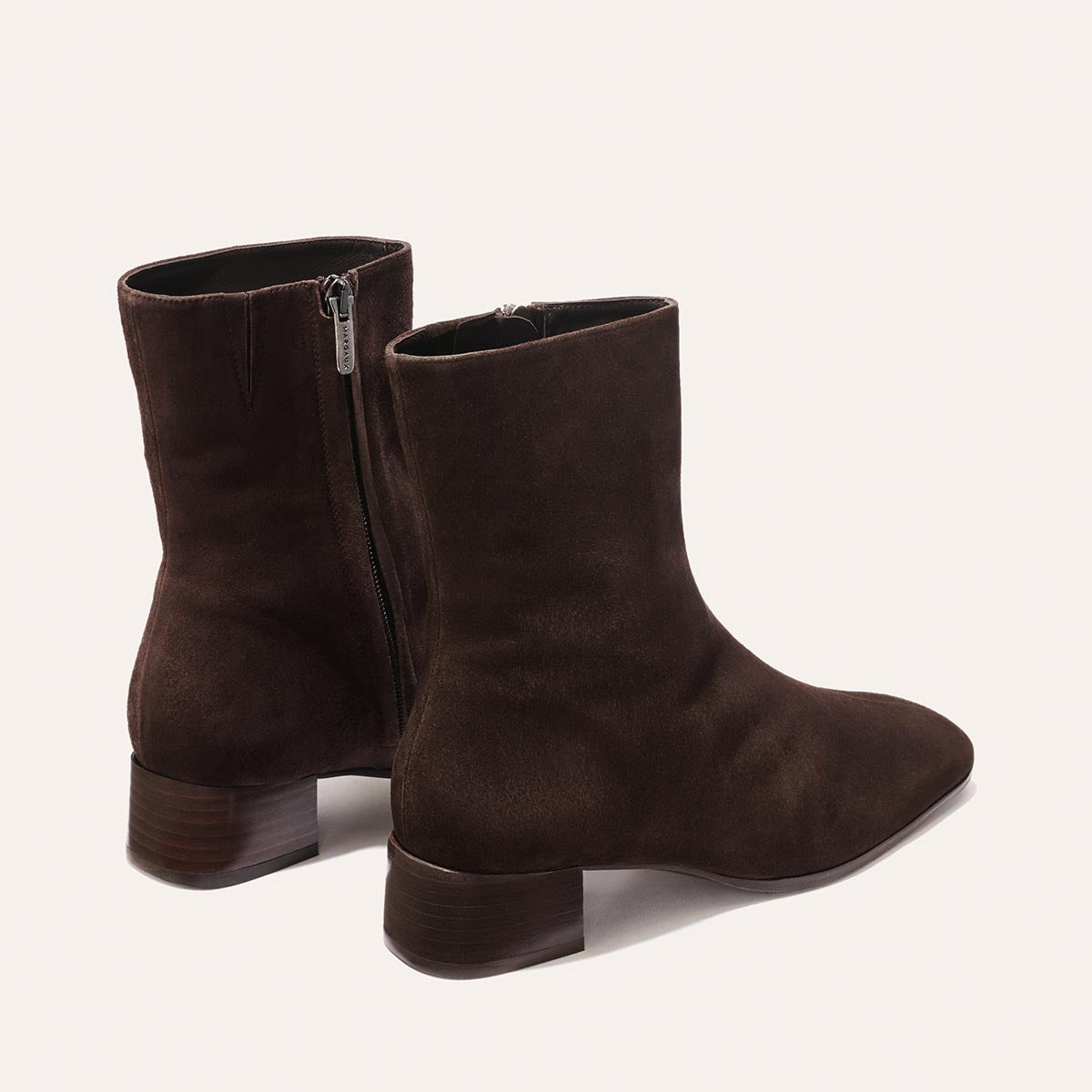 The Phoebe Boot - Molasses Suede by Margaux is a pair of dark brown suede ankle boots with side zippers and low wooden heels, expertly crafted using the artisanal sacchetto technique for exceptional flexibility and finished with a padded insole for added comfort, shown against a plain light background.
