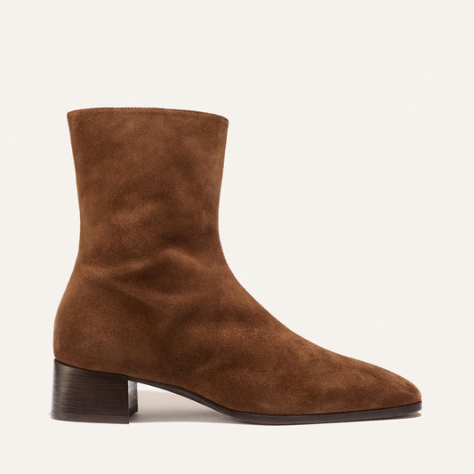 Introducing The Phoebe Boot - Ganache Suede by Margaux: a brown suede ankle boot featuring a rounded toe, low block heel, and minimalist silhouette—expertly crafted with the artisanal sacchetto technique for superior flexibility and comfort. Presented in side profile against a plain light background.