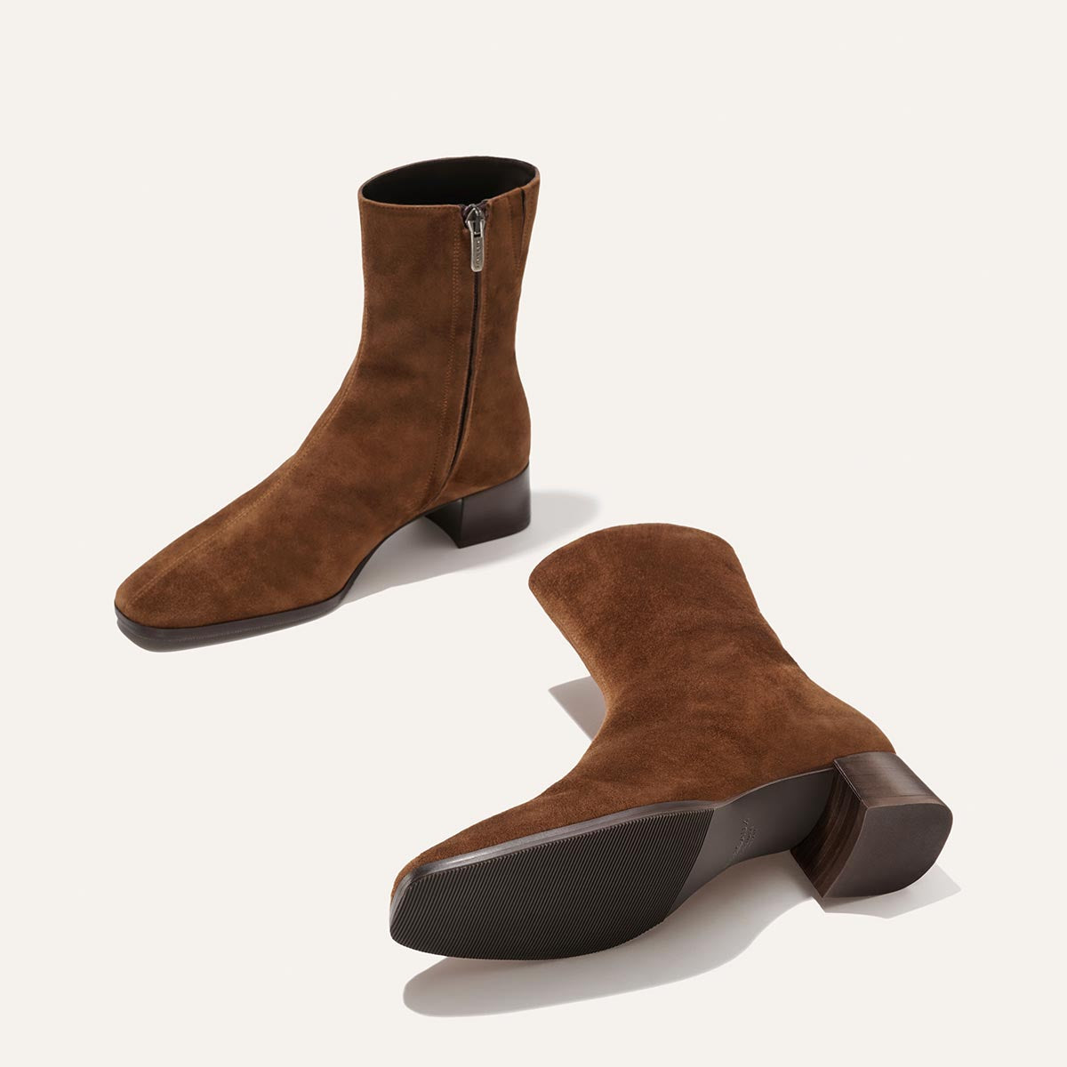 A pair of The Phoebe Boot – Ganache Suede by Margaux, featuring rich brown suede, low block heels, and side zippers, expertly crafted with the artisanal sacchetto technique for superior flexibility; one boot is standing upright while the other lies on its side against a plain light background.