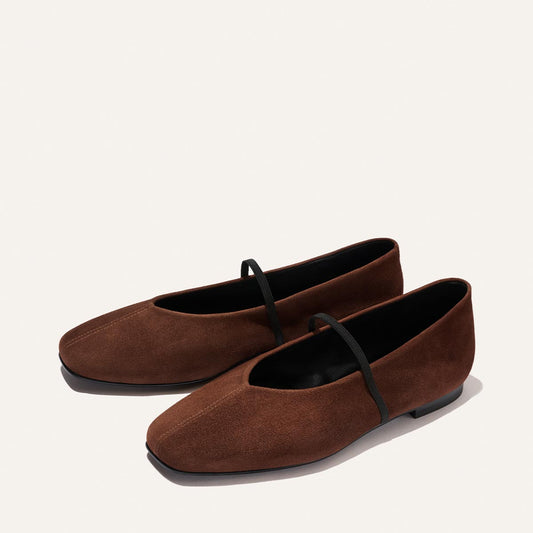A pair of Margaux's The Phoebe - Ganache Suede ballet flats, featuring rich brown suede, black soles, and a single black elastic strap across each shoe, expertly crafted with sacchetto construction for outstanding flexibility and comfort, displayed against a light background.