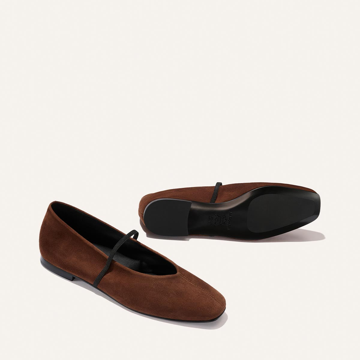 The Phoebe - Ganache Suede by Margaux: a pair of brown suede ballet flats with black soles and slim black straps, one placed upright and the other lying on its side against a plain light background. Expertly crafted with sacchetto construction, these could be the softest flats you'll ever wear.