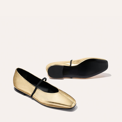 The Phoebe - Gold Crinkle Nappa by margaux-development: gold ballet flats with black soles and a thin black strap, crafted using sacchetto construction for ultimate softness. One shoe stands upright, the other shows its sole against a light background.
