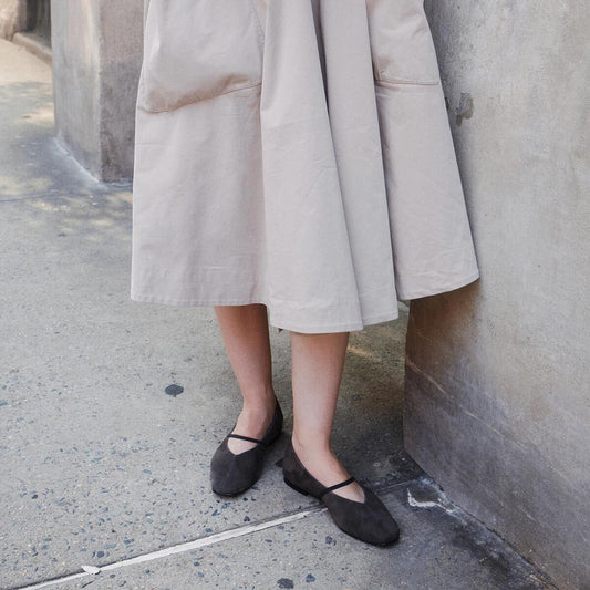 A person in a light beige skirt stands by a concrete wall, wearing Margaux’s The Phoebe - Graphite Suede flats—crafted with sacchetto construction for an exceptionally soft fit—on a sidewalk.
