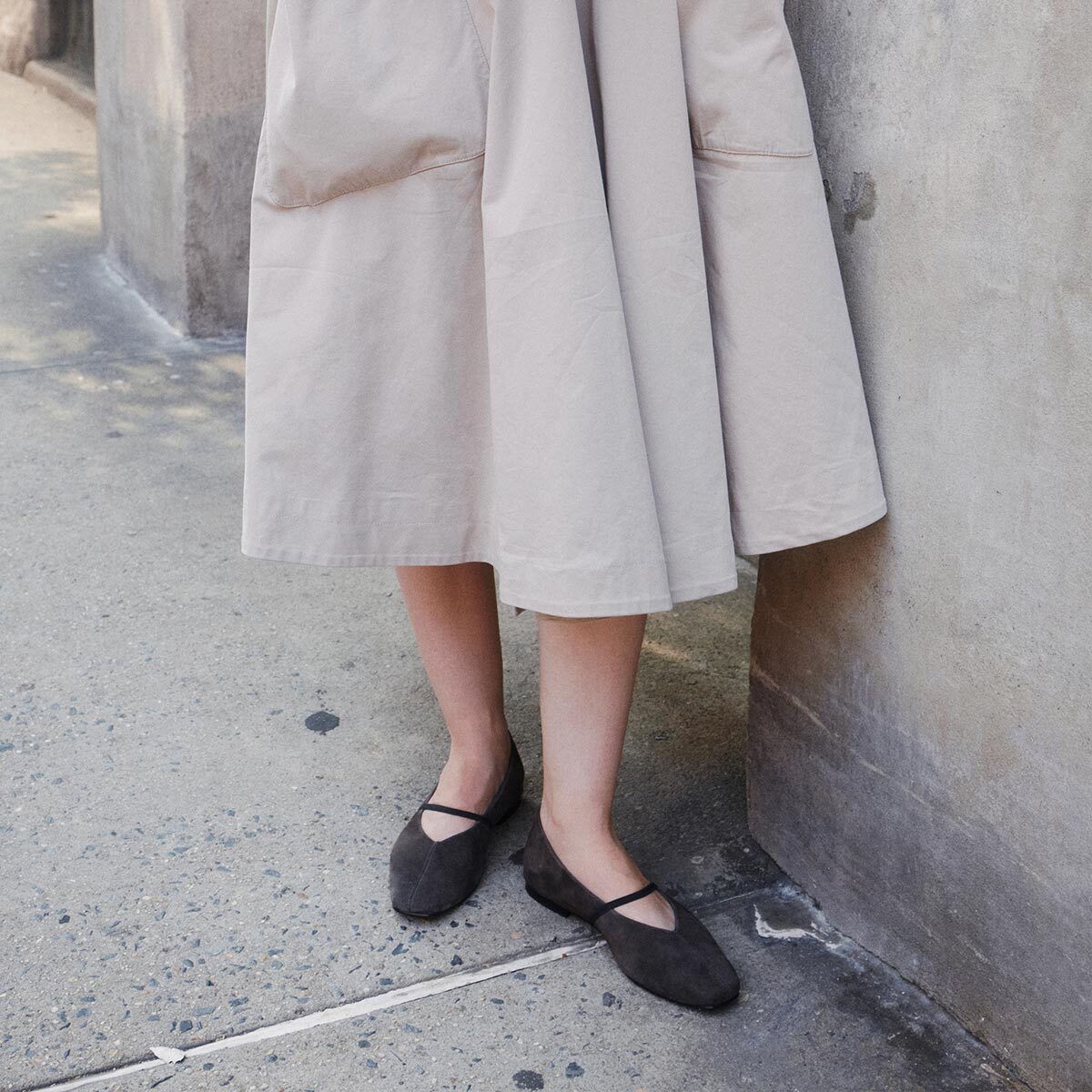 A person in a light beige skirt stands by a concrete wall, wearing Margaux’s The Phoebe - Graphite Suede flats—crafted with sacchetto construction for an exceptionally soft fit—on a sidewalk.