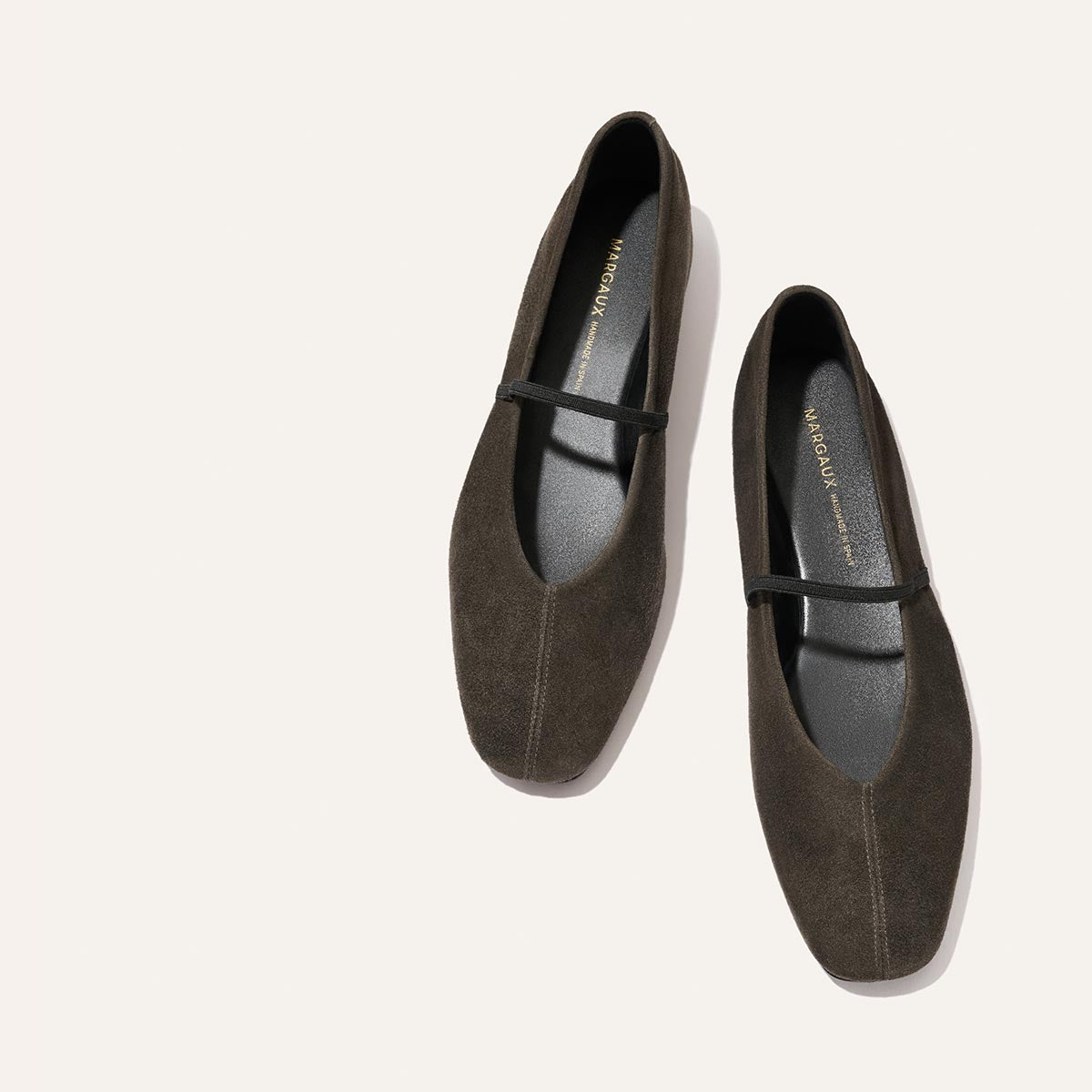 A pair of The Phoebe - Graphite Suede Mary Jane flats by Margaux, beautifully crafted with sacchetto construction for outstanding comfort, featuring black insoles and a slim strap across the top, displayed against a light beige background.
