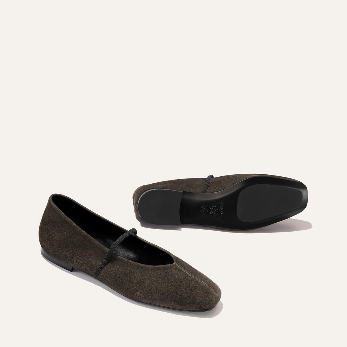 A pair of The Phoebe - Graphite Suede Mary Jane flats by Margaux, handmade with sacchetto construction for an ultra-soft feel, shown with one shoe facing up and the other on its side, both featuring graphite suede uppers, black soles, and a delicate strap across the top.