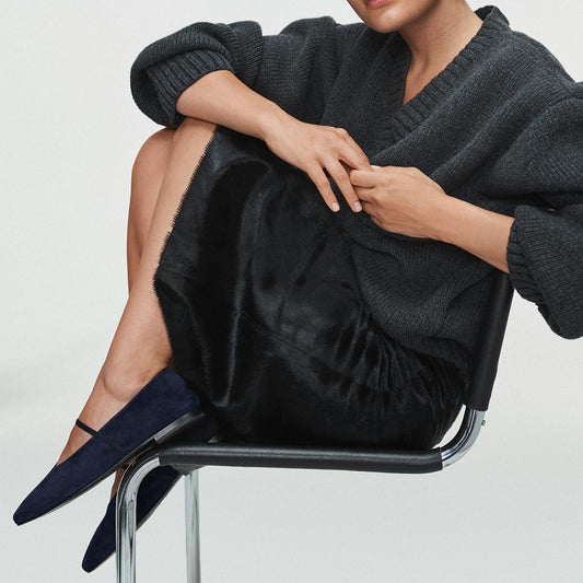 A person in a dark sweater and black skirt sits sideways on a chair, legs crossed and hands resting on their lap, wearing Margaux’s The Phoebe flats in indigo suede—pointed-toe shoes crafted with sacchetto construction for the softest fit.