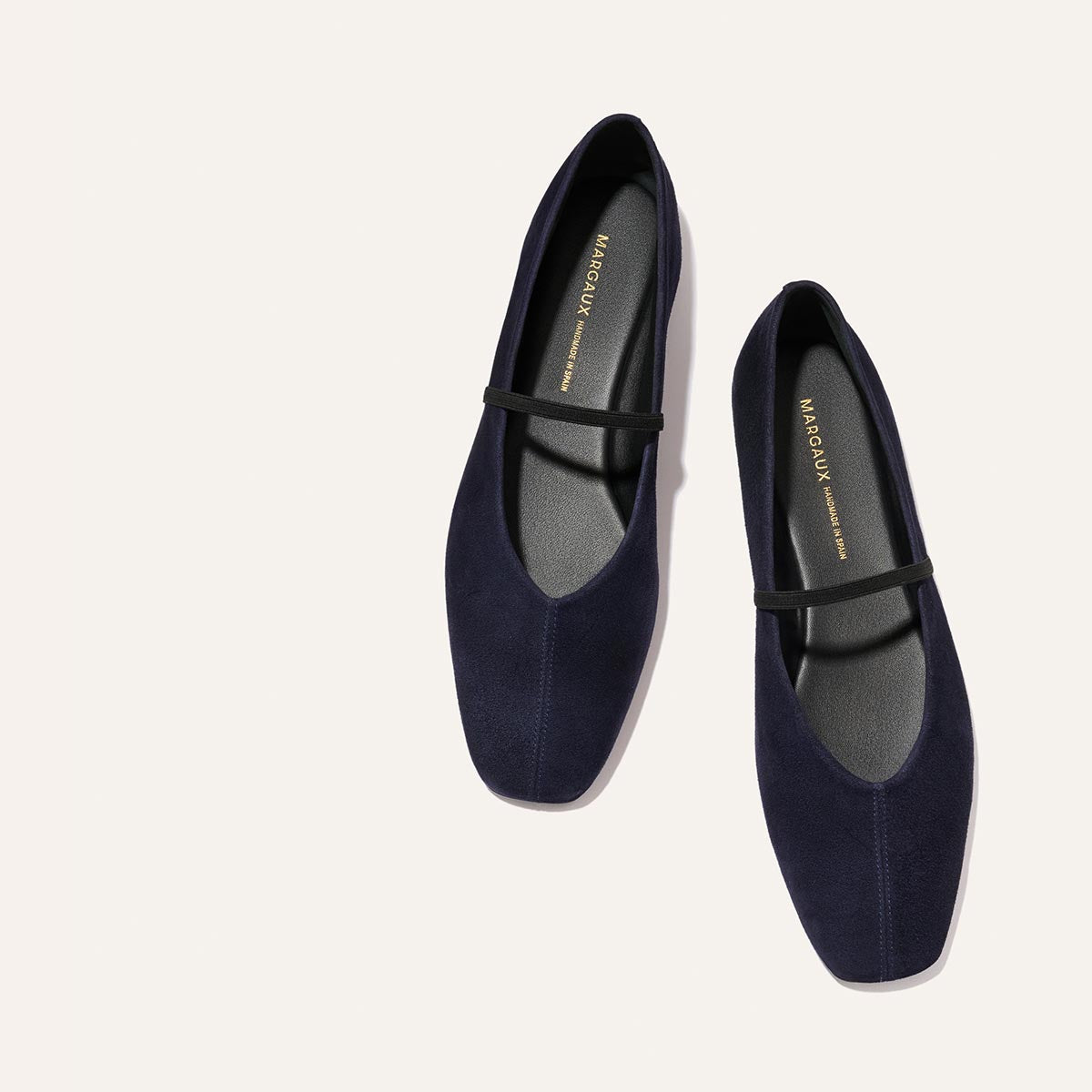 A pair of The Phoebe flats in indigo suede by Margaux, featuring black straps, square toes, and the brand’s gold logo on black insoles, displayed on a light background. Crafted with sacchetto construction for remarkable flexibility, these are truly the softest flats you’ll slip into.