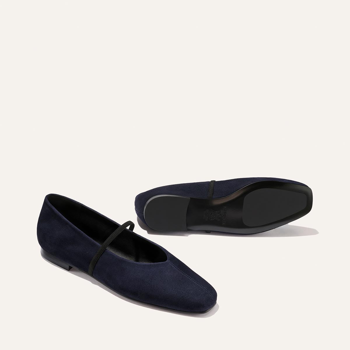 Two Indigo Suede "The Phoebe" ballet flats by Margaux, featuring black soles and a slender black strap, expertly crafted with sacchetto construction for outstanding flexibility and comfort, are displayed against a plain light background—one upright and the other sideways to reveal the sole.