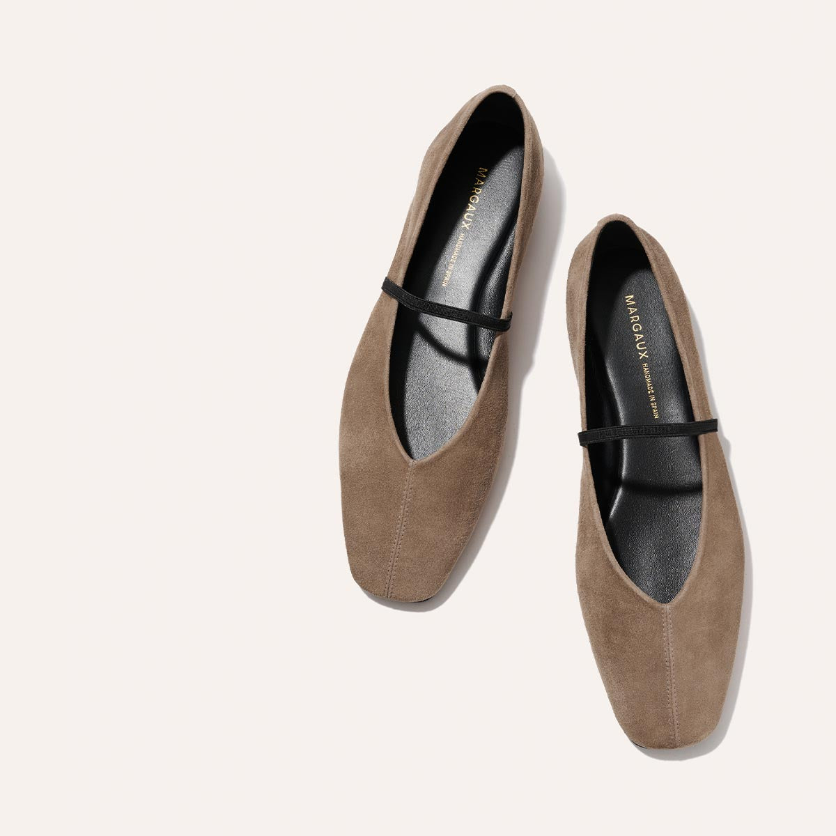 The Margaux Phoebe – Mushroom Suede flats feature taupe suede, black top straps, and a slightly squared toe, shown on a light background.