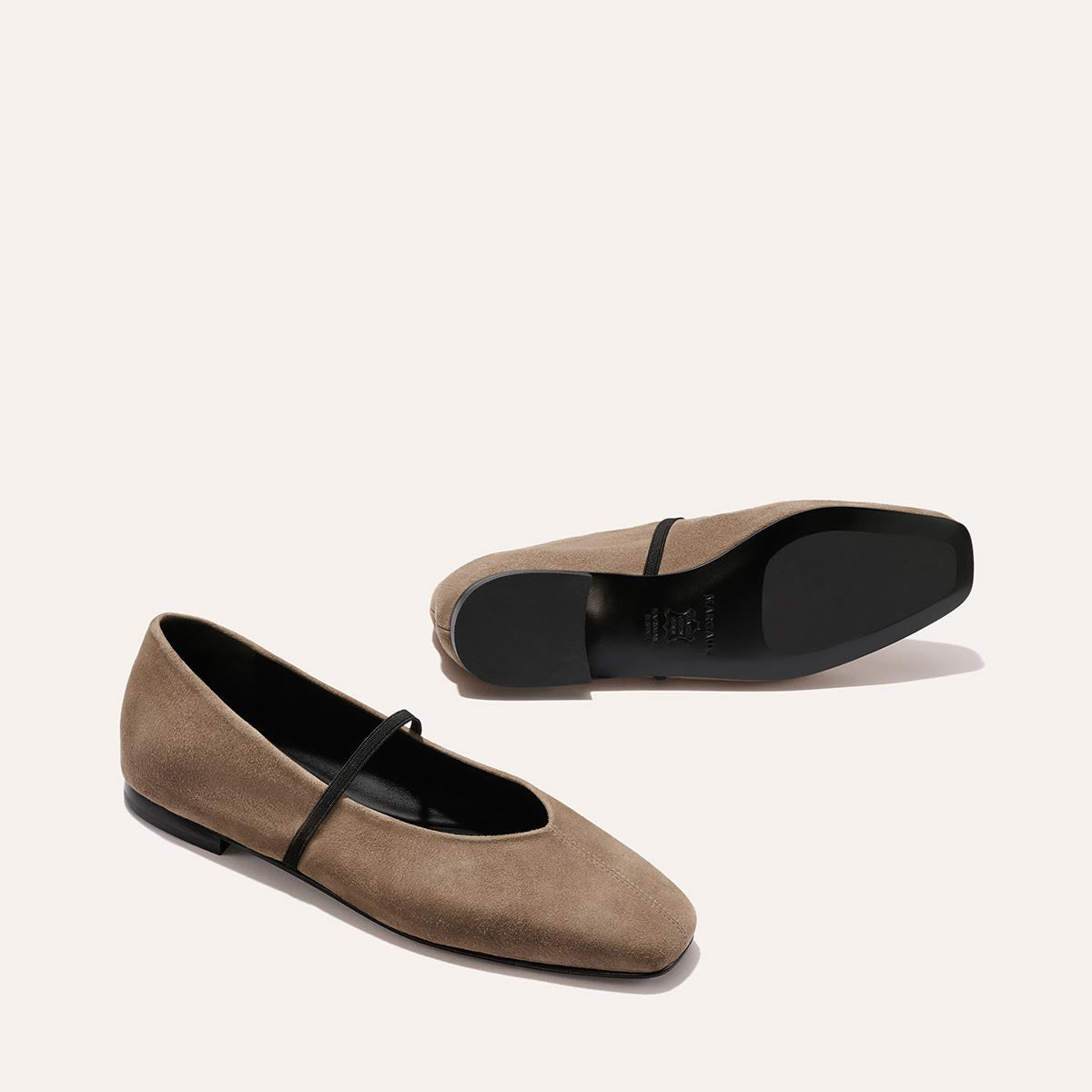 A pair of Margaux's The Phoebe - Mushroom Suede Mary Jane flats with a black strap, one flat and one on its side displaying the sole, set against a plain off-white background.