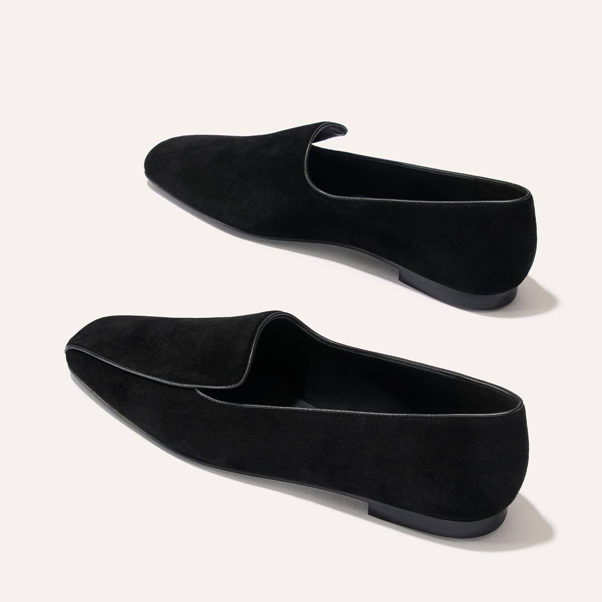 A pair of The Portia Loafers in Black Suede by Margaux, featuring a slight heel and showcased on a light background. The shoes display a simple, elegant design with subtle trim along the edges.