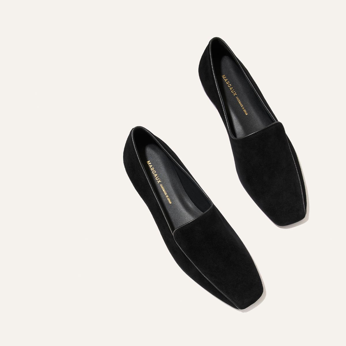 The Portia Loafer in black suede by Margaux is shown from above on a light background, featuring square toes and gold branding on the insole.