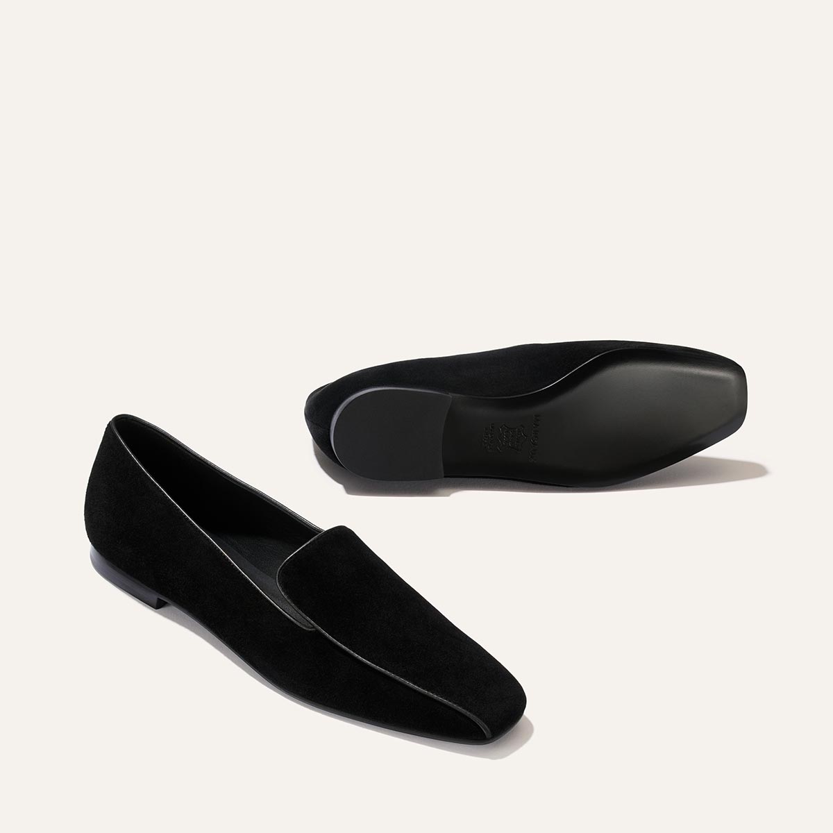 The Portia Loafer in Black Suede by Margaux is displayed, with one shoe standing upright and the other positioned on its side to showcase the sole.