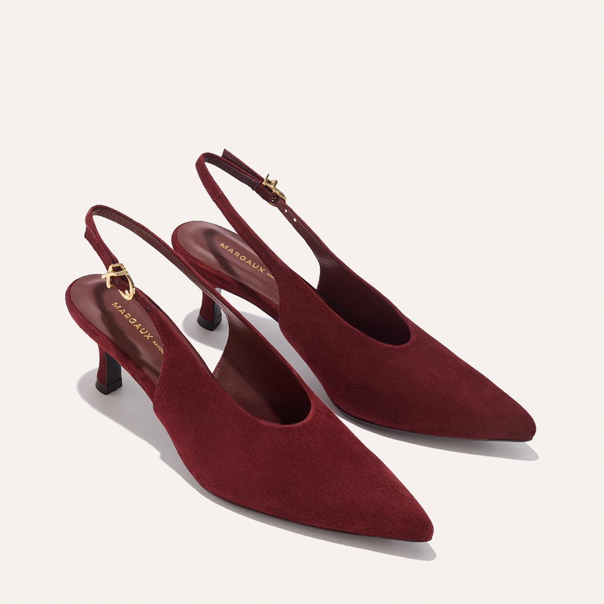 The Prado Sling - Burgundy Suede by margaux-development features elegant pointed-toe slingback heels in deep burgundy suede with low heels and gold buckle accents on the ankle straps, displayed on a light background.