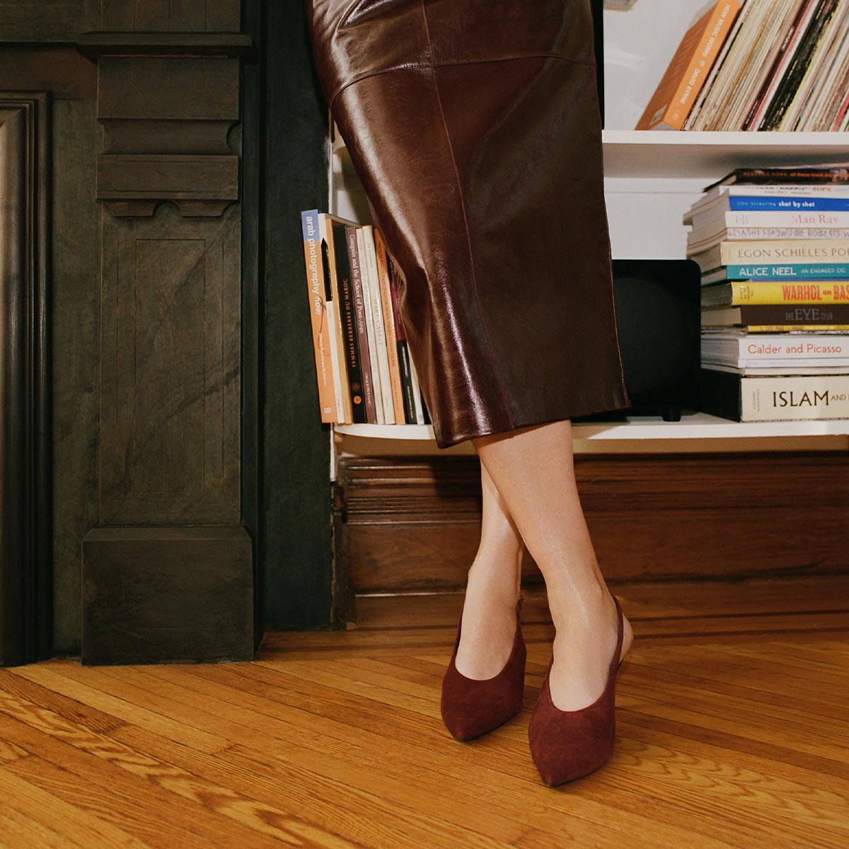 Wearing a brown leather skirt and The Prado Sling - Burgundy Suede by margaux-development, a person stands on a wooden floor, leaning against a fireplace near a bookshelf filled with books. Their upper body is not visible.