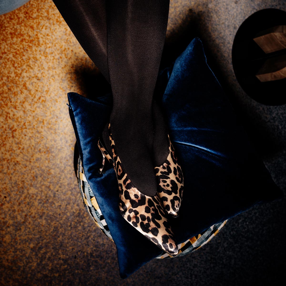 A person wearing black tights and The Prado Sling - Leopard Satin by margaux-development rests their feet on a blue velvet cushion, with a textured surface and a hat partially visible in the background.