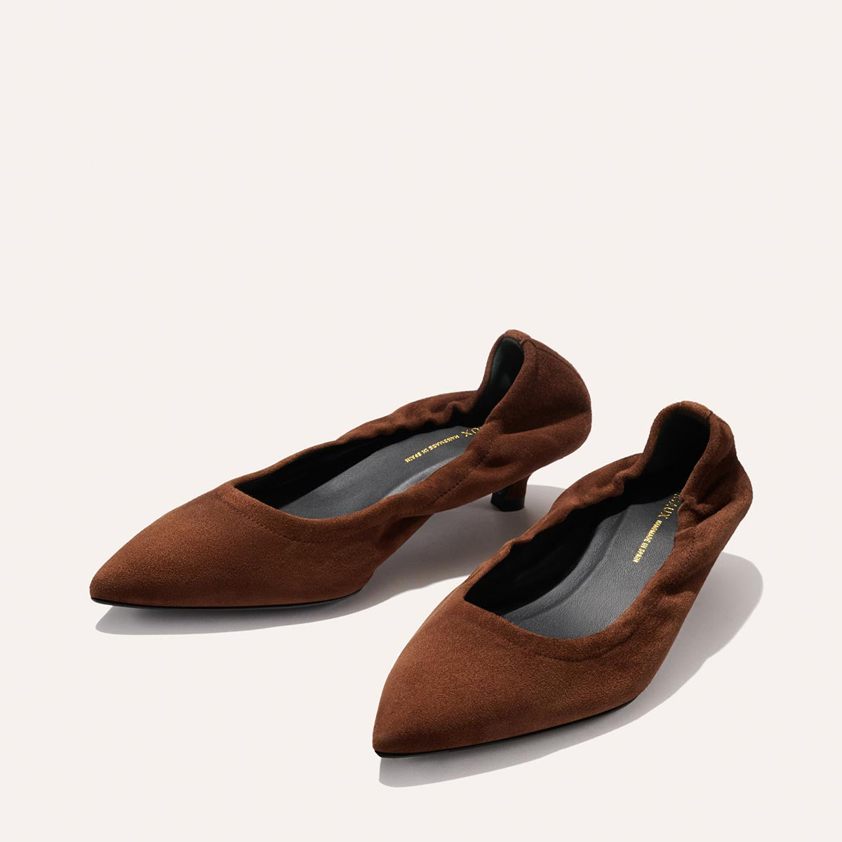 A pair of The Roma 40 - Ganache Suede heels by Margaux, crafted in brown suede with ruffled edges, pointed toes, a comfortable walkable heel, and black insoles—elegantly displayed against a plain light background.
