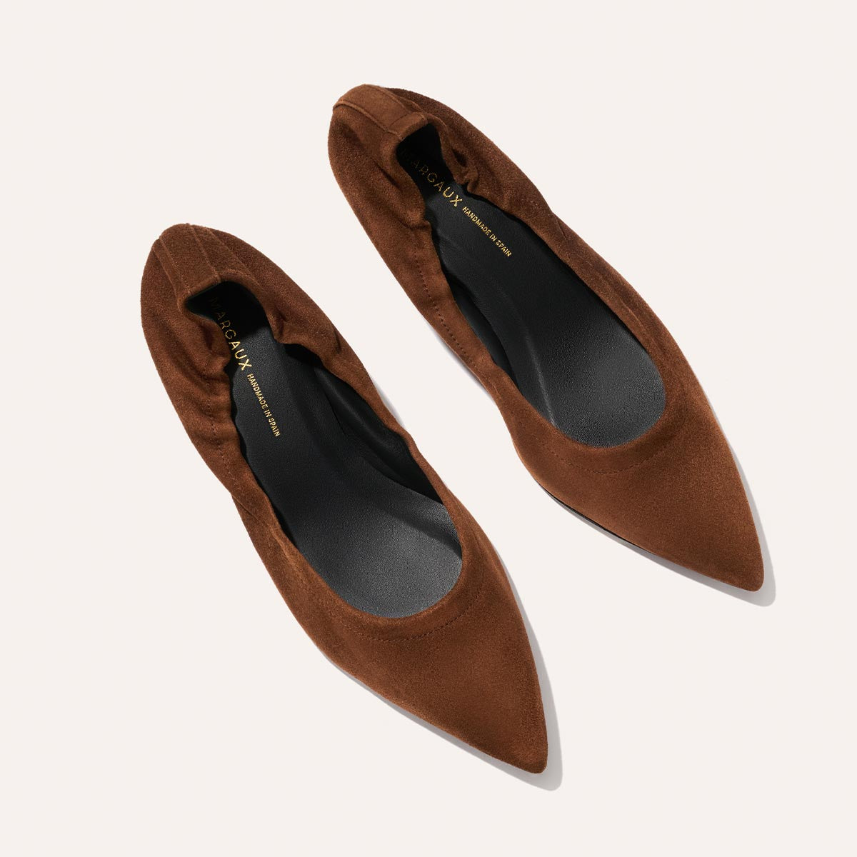 A pair of Margaux The Roma 40 - Ganache Suede flats, in brown suede-like material with black cushioned insoles and subtle ruching at the edges, shown from above on a light background.