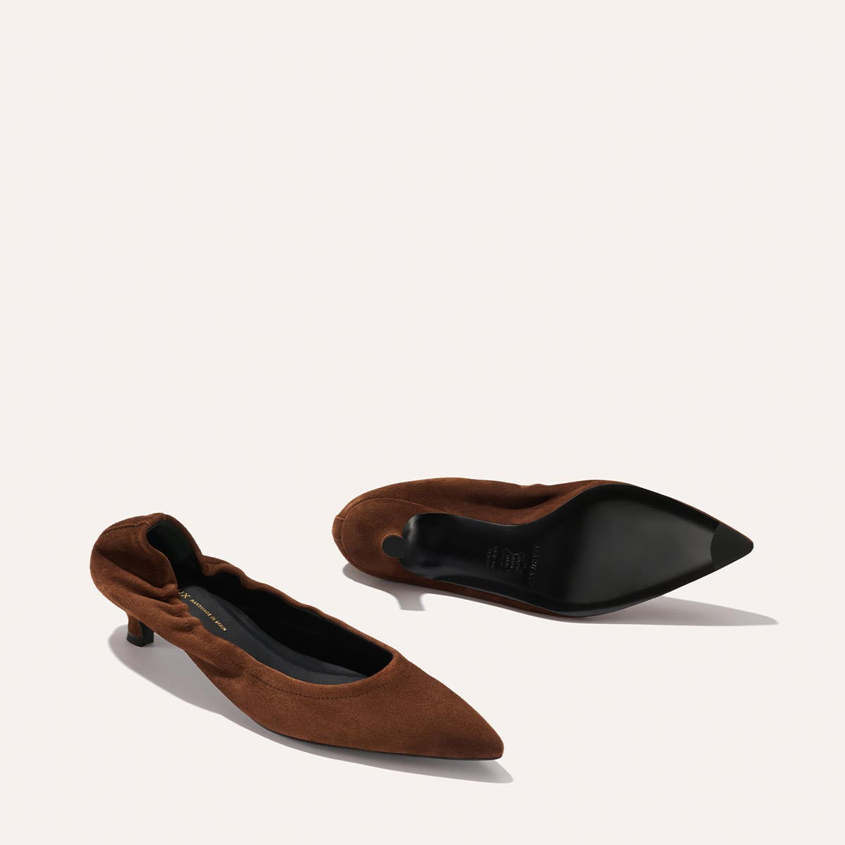 The Roma 40 - Ganache Suede by Margaux is shown: one brown suede kitten heel upright, the other on its side to reveal the black sole. These pointed-toe heels feature elegant ruching along the edges.