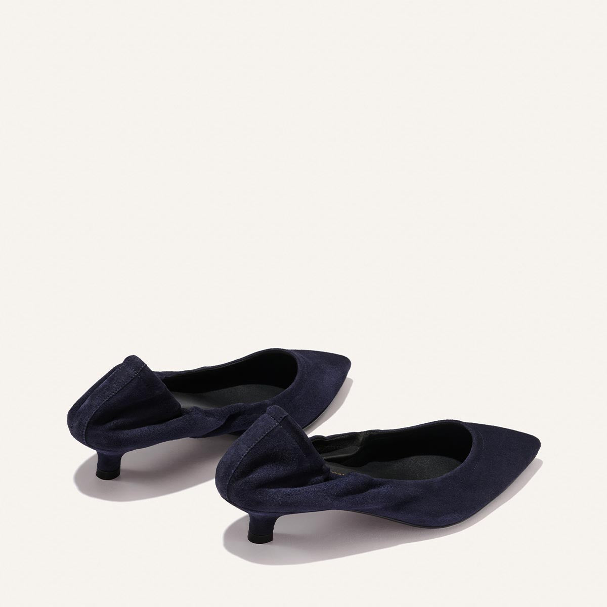 Two indigo suede pointed-toe kitten heel shoes, known as The Roma 40 by Margaux, feature a soft fabric bow detail on the heel and offer a comfortably walkable height. They are displayed against a plain off-white background.