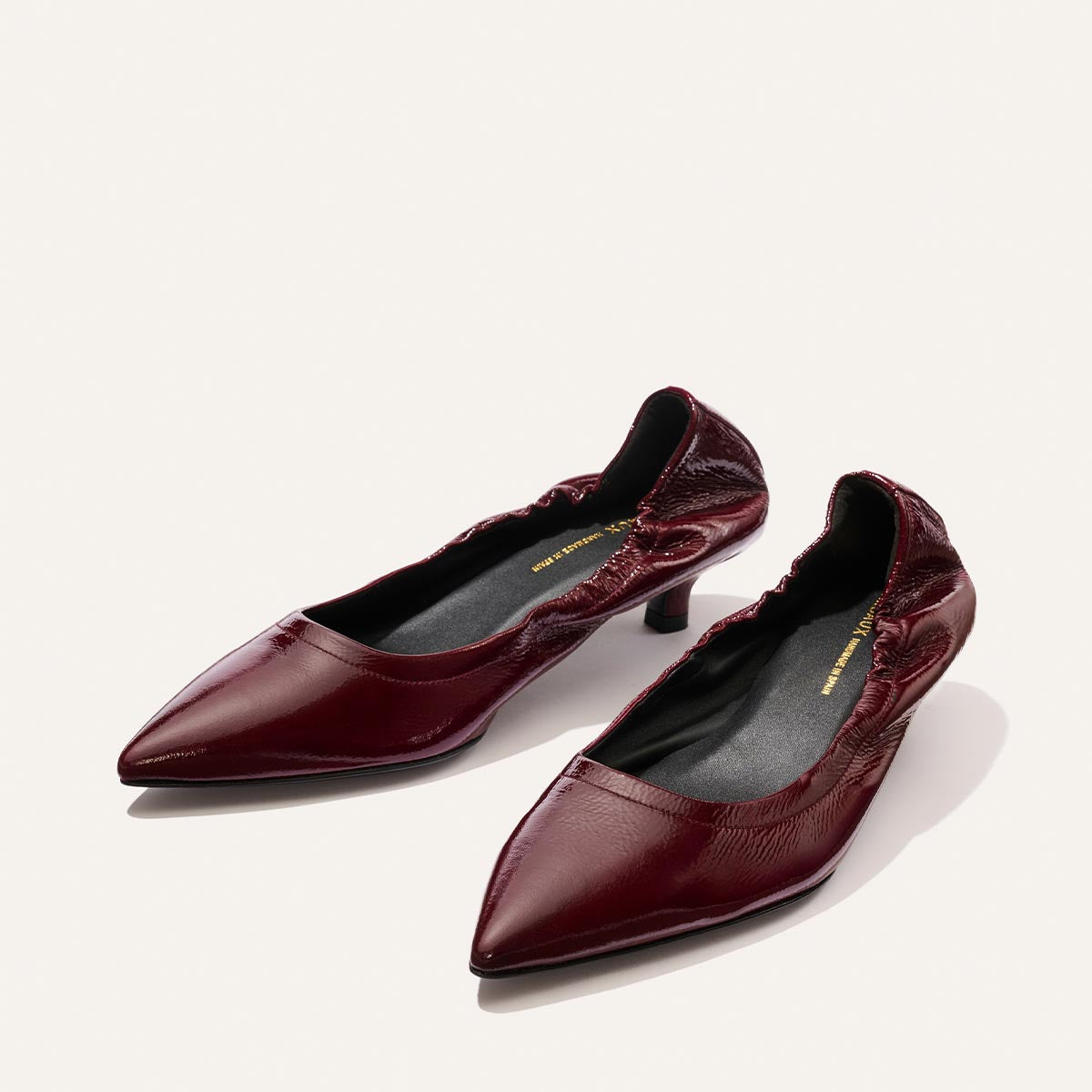 The Margaux Roma 40 - Merlot Crinkle Patent is a pair of burgundy pointed-toe flats crafted from patent leather, featuring a low, walkable heel and gathered detailing around the opening, displayed against a light background.