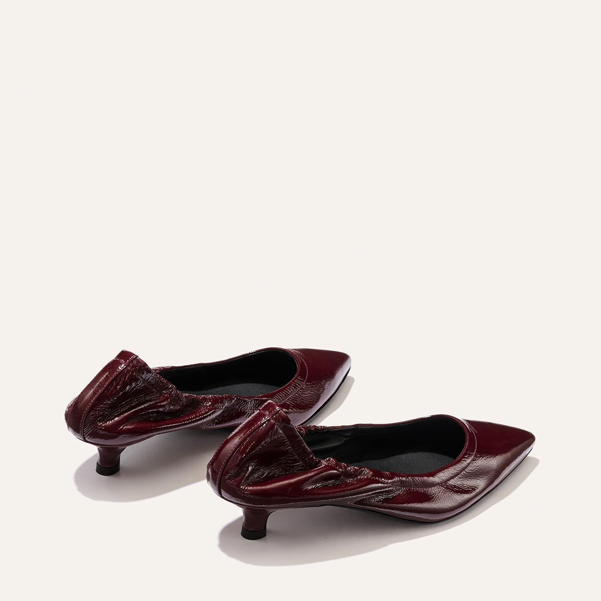 A pair of Margaux's The Roma 40 shoes in Merlot Crinkle Patent are displayed on a plain light background. These glossy burgundy low-heeled shoes feature pointed toes and gathered detailing at the heel, blending the elegance of a pointed-toe flat with the comfort of a walkable heel.