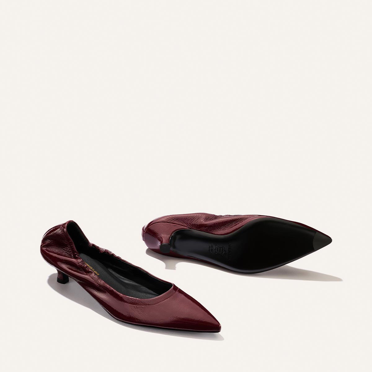A pair of Margaux’s The Roma 40 - Merlot Crinkle Patent shoes—one upright and one resting on its side—are displayed against a simple light background. Their walkable heel offers comfort while maintaining a chic, pointed-toe silhouette.