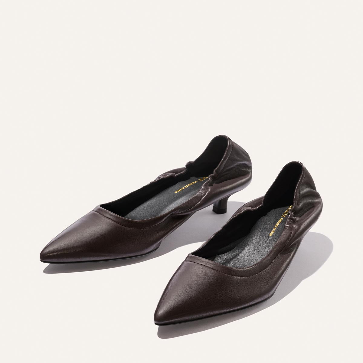 A pair of espresso nappa, pointed-toe flat women’s shoes by Margaux, featuring a slightly ruched design near the opening and a chic heel, displayed on a light background—The Roma 40 effortlessly blends elegance with comfort.
