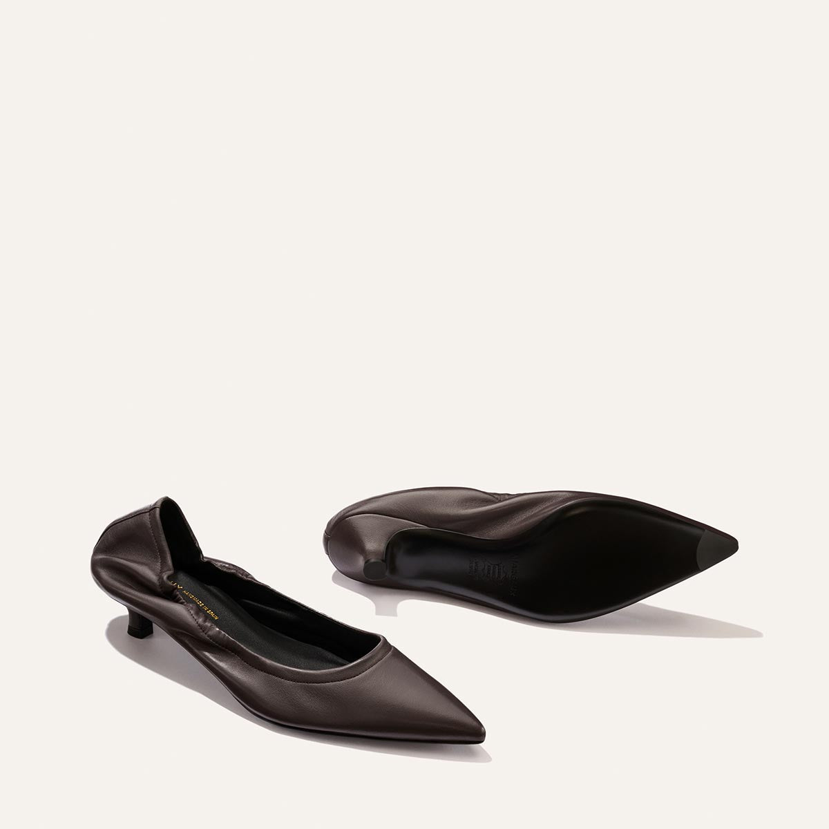 A pair of Margaux's The Roma 40 - Espresso Nappa pointed-toe kitten heel shoes is displayed on a light background, with one elegant heel standing upright and the other positioned on its side to showcase the sole.