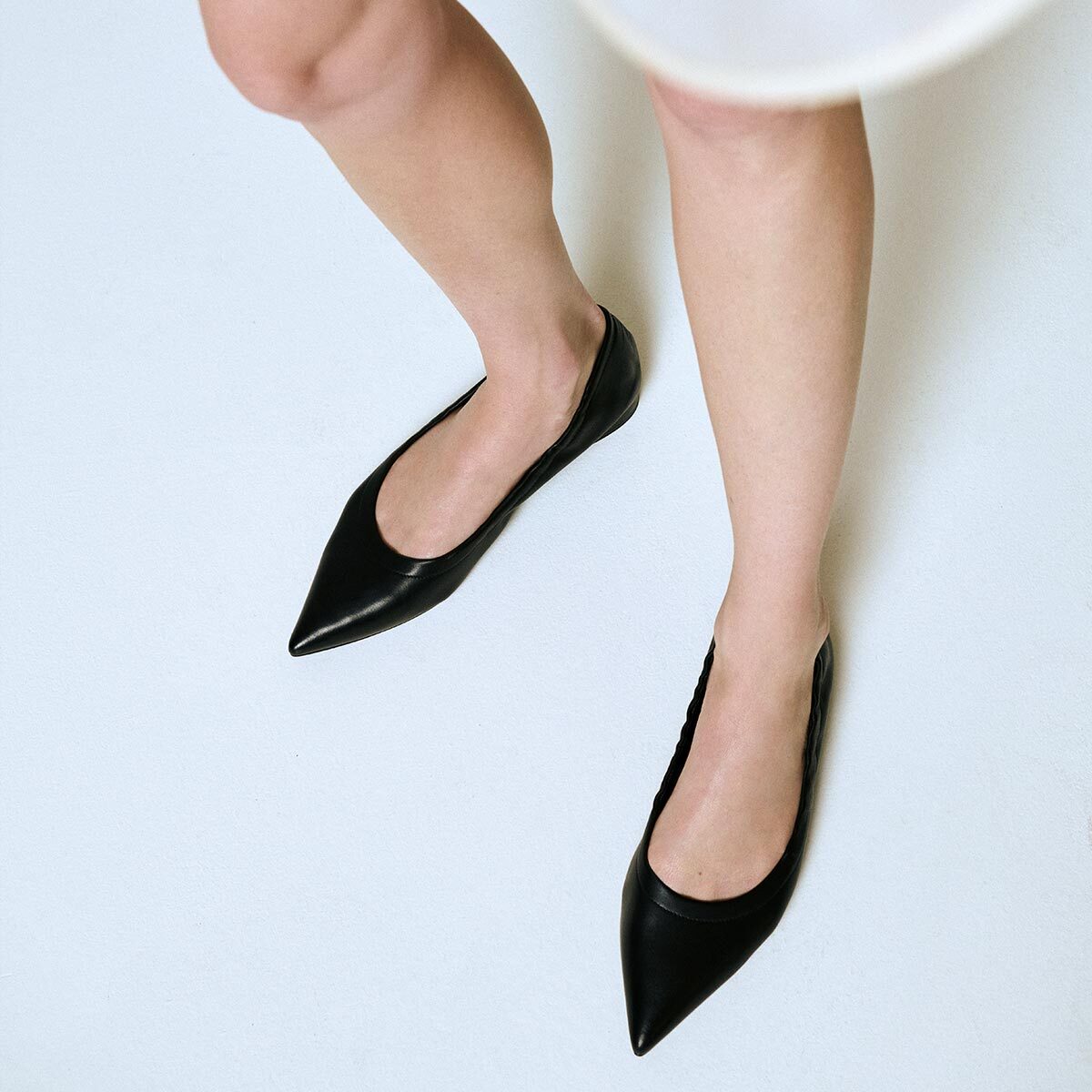 A person stands on a white surface, wearing a white skirt and Margaux’s The Roma - Black Nappa pointed-toe ballet flats. The image, shot from above, highlights the sophisticated footwear and legs.
